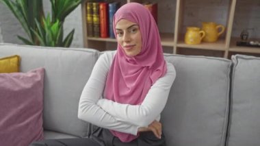 Parlak gözlü, tesettürlü, tebessümlü, koltukta kendinden emin bir şekilde oturan İspanyol bir kadın. Pozitif titreşimler yayıyor, kollarını kavuşturuyor, evinin rahat ortamından sana bakıyor..