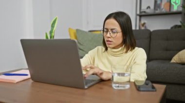 Modern bir oturma odasında dizüstü bilgisayar kullanan, gözlüklü, konsantre olmuş genç İspanyol kadın kafa karışıklığı ve hayal kırıklığı ifadeleri sergiliyor..