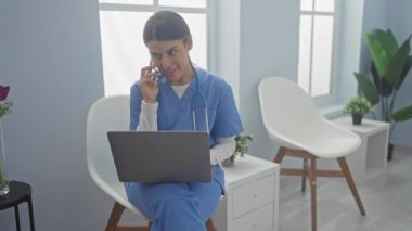 Mavi önlüklü genç bir kadın telefondayken modern bir klinikte dizüstü bilgisayarla çalışıyor..