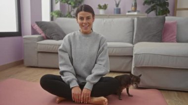 Sıcak bir oturma odasında yoga minderinin üzerinde Chihuahua 'sıyla bacak bacak üstüne atmış gülümseyen bir kadın..