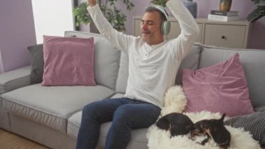 Orta yaşlı bir adam rahat bir kanepede chihuahualarla dinleniyor, kulaklık takıyor ve modern bir oturma odasında müziğin tadını çıkarıyor..