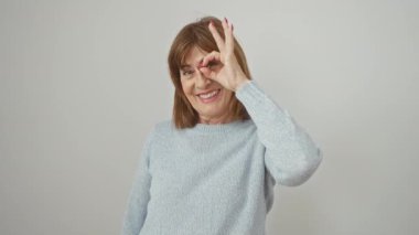Süveter giyen neşeli, orta yaşlı bir kadın, 'ok' jesti, parmak dürbününden röntgenci, geniş bir gülümseme. Hepsi temiz ve izole beyaz bir arka planda..