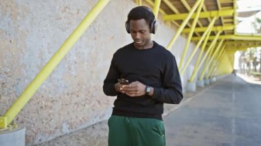 Kulaklıklı Afro-Amerikalı bir adam şehrin caddesinde ayakta ve koşarken akıllı telefon kullanıyor.