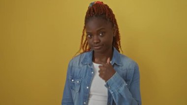 Zarif Afro-Amerikalı kadın kot gömlek giyiyor, izole edilmiş sarı arka planda duruyor, 'seni izliyorum' jestinde gözüne işaret ediyor.