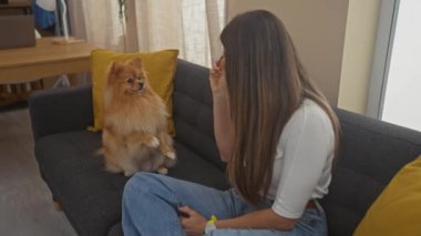 Genç bir kadın rahat bir dairede kanepede Pomeranian köpeğiyle oynuyor.