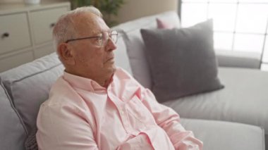 Yaşlı gri saçlı adam oturma odasında oturuyor, gözlüklü ve pembe gömlekli, yüzünde düşünceli bir ifade var..