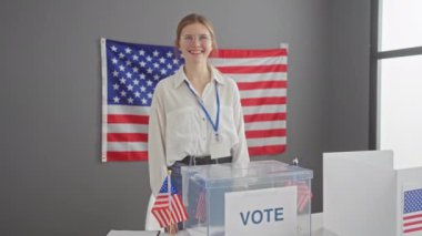 Gözlüklü, gülümseyen, beyaz bir kadın, arka planda Amerikan bayrağı olan bir seçim merkezinde başparmak kaldırıyor..