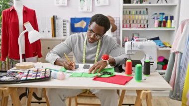 Manken üzerinde kırmızı kumaşı olan iyi donanımlı bir atölyede terzilik yapan Afrikalı bir adam renkli iplikler arasında çizimler yapıyor..