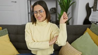 Neşeli genç İspanyol kadın evde, neşeyle yan tarafı gösteriyor, güvenle gözlüklerinin arkasında gülümsüyor.