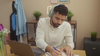 Evde sakallı, kulaklık takan yakışıklı latin adam işe odaklanmış notlar yazıyor.