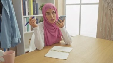 Pembe tesettürlü genç bir kadın elinde kalemle akıllı bir telefona konuşuyor, ofis masasında oturuyor..