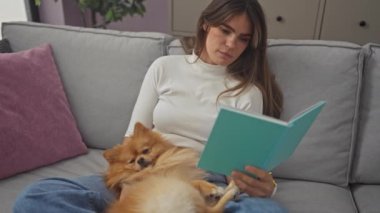Genç bir kadın, evinde kanepede kitap okumaktan zevk alır. Yanında Pomeranian köpeği ile..