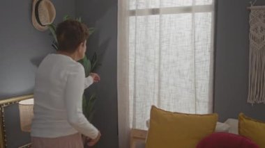 Kısa saçlı yaşlı bir kadın yatak odasının penceresinden içeri bakar. Etrafı iç mekan bitkileriyle ve dekorla çevrilidir..