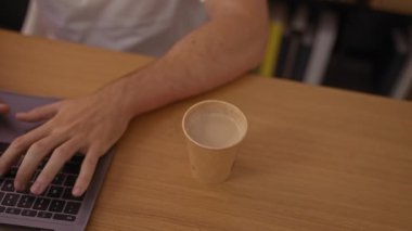 Organize bir ofiste ya da kapalı bir iş yerinde dizüstü bilgisayarda çalışan genç bir adam, masada içecek bardağıyla teknoloji kullanıyor..