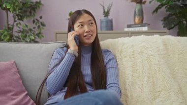 Genç Çinli bir kadın, evdeki güzel dekore edilmiş oturma odasında rahat bir kanepede otururken akıllı bir telefonla konuşuyor..
