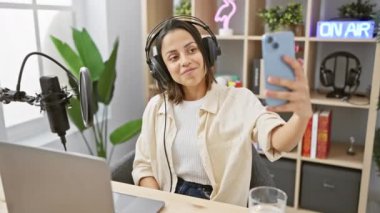 Genç İspanyol kadın, modern bir radyo stüdyosunda podcast kaydında özçekim yapıyor. Çekici gülüşünü ve saç stilini sergiliyor..