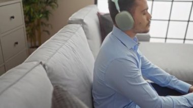Genç İspanyol adam büyük kulaklıklarla müzik dinliyor, modern oturma odasındaki gri bir kanepede dinleniyor..