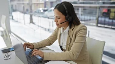 Kulaklığı olan genç, İspanyol bir kadın, parlak, modern bir ofis alanında dizüstü bilgisayarla çalışıyor, profesyonellik yayıyor..
