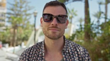 Güler yüzlü, dövmeli bir adam, güneşli bir parkta, bulanık şehir geçmişiyle güneş gözlüğünü çıkarıyor..