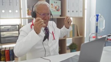 Klinikteki yaşlı bir doktor kulaklık ve steteskop takıyor, gülümsüyor ve tıbbi ekipman ve ofis malzemeleriyle çevrili bir laptopla iletişim kuruyor..