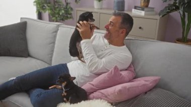Orta yaşlı İspanyol bir adam oturma odasında rahat bir kanepede iki chihuahua köpeğiyle oynuyor. Etrafı rahat dekor ve ev bitkileriyle çevrili..
