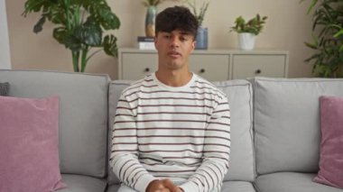 Modern bir oturma odasında gri bir kanepede oturan genç İspanyol bir adam saksı bitkileri, dekoratif yastıklar ve çizgili beyaz bir gömlek giyen küçük bir dolap..