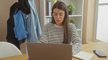 Modern bir ev ofisinde dizüstü bilgisayarla çalışan, kahve ve akıllı telefonlu İspanyol bir kadın..
