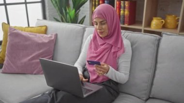 Evde bir kanepede dizüstü bilgisayar ve kredi kartı kullanan tesettürlü gülümseyen bir kadın online alışveriş veya bankacılık tasvir ediyor..