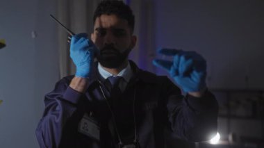 Sakallı, eldivenli ve dedektif rozetli odaklanmış bir adam kapalı bir suç mahallinde telsizle iletişim kuruyor..