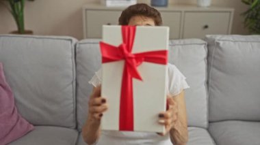 Kısa saçlı, yaşlı bir İspanyol kadın, parlak bir oturma odasında kırmızı kurdeleli bir hediye tutuyor ve kapalı alanda neşeli bir kutlama yapıyor..