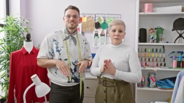 Bir erkek ve kadın moda tasarımcıları, manken ve aletlerle donanımlı bir terzi dükkanında iş birliği yapıyorlar.