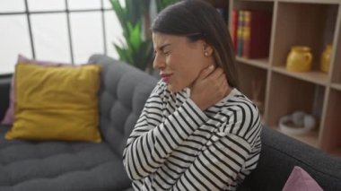 Modern bir oturma odasındaki gri koltukta oturan boynu ağrıyan İspanyol kadın.