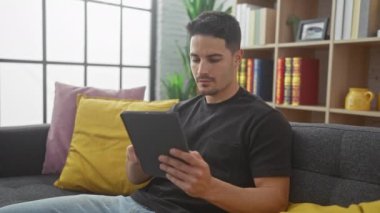 Gündelik oturma odasındaki yakışıklı İspanyol bir adam odaklanmış bir tablet kullanıyor, teknolojiyi ve yaşam tarzını aktarıyor..