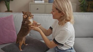 Genç sarışın bir kadın, sıcak ve sevgi dolu bir ev ortamı sergileyerek, rahat bir oturma odasında küçük chihuahua köpeğiyle etkileşime geçip oynuyor..