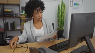 Genç, güzel, kıvırcık saçlı Afro-Amerikalı bir kadın ofiste oturmuş, ofis malzemeleriyle çevrili ve masasında bir bilgisayar ile gözlüğünü düzeltirken bir belge okuyor..