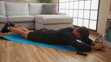 Genç bir İspanyol adam evdeki mavi bir paspasın üzerinde egzersiz yapıyor, gri bir kanepe ve pencerenin yanındaki ahşap zeminde pilates yapıyor..