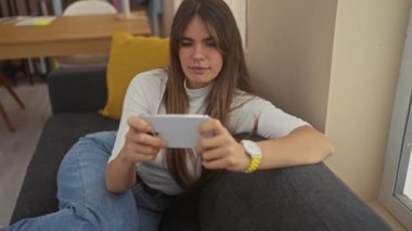 Gülümseyen genç bir İspanyol kadın, iyi ışıklandırılmış oturma odasındaki gri bir kanepede otururken akıllı telefonuyla video oyunu oynuyor..