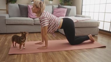 Sarışın genç bir kadın oturma odasındaki pembe bir paspasın üzerinde yoga yapıyor. Yanında bir Chihuahua köpeği var. Rahat bir kanepe ve kapalı salonda bitkiler sergiliyor..