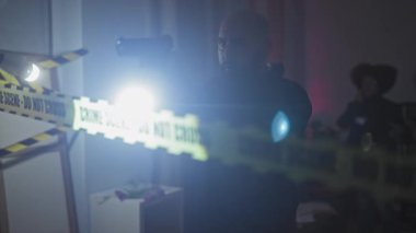 İçerideki bir suç mahallini iki ajan araştırıyor. Bir adam silah doğrultuyor ve bir kadın telefon tutuyor..