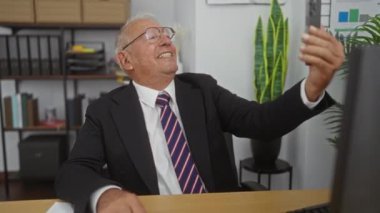 Beyaz saçlı, beyaz takım elbiseli yaşlı bir adam modern bir ofiste arka planda raflar ve bitkilerle selfie çekiyor..