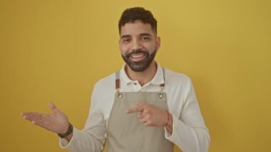 Genç İspanyol adam şaşırmış ve gülümsüyor, önlük giyiyor, elini uzatıyor ve işaret ediyor, izole edilmiş sarı bir arka planın başında duruyor..