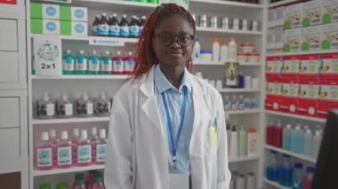 İyi stoklanmış bir eczanenin içinde beyaz laboratuvar önlüğüyle gülümseyen genç Afrikalı Amerikalı eczacı.