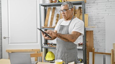 Mature man examines a tablet in his woodshop filled with various carpentry tools. clipart
