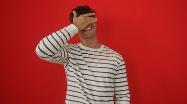 Yakışıklı, genç, İspanyol bir adam, çizgili bir süveter giyerken izole edilmiş kırmızı bir arka plana karşı poz vererek tek eliyle gözlerini kapatıyor..