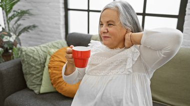 Son sınıf öğrencisi bir kadın, rahat bir oturma odasında, kanepede kahve fincanıyla rahatlıyor.