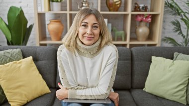 Kafkasyalı yetişkin bir kadın, sarı saçlı rahat bir oturma odasında gülümseyen, dost canlısı ve olgun bir aura sergiliyor..