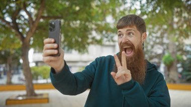 Sakallı bir adam parkta selfie çeker, barış işareti yapar, dışarıda neşe ifade eder..