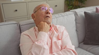 Pembe tişörtlü yaşlı, gri saçlı adam oturma odasındaki kanepede, sakin bir evde, düşünceli bir şekilde oturur..
