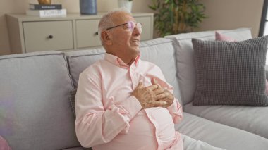 Evdeki kanepede oturan yaşlı bir adam, gözlük takıyor ve göğsünü tutuyor. Bu da oturma odasındaki kalp sorunlarını gösteriyor..