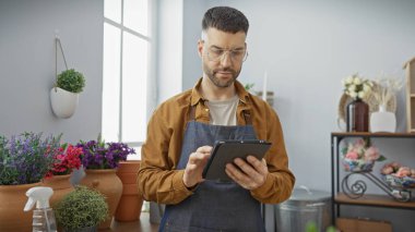Kapalı bir çiçekçide renkli çiçeklerin arasında tablet kullanan önlüklü yakışıklı İspanyol bir adam..
